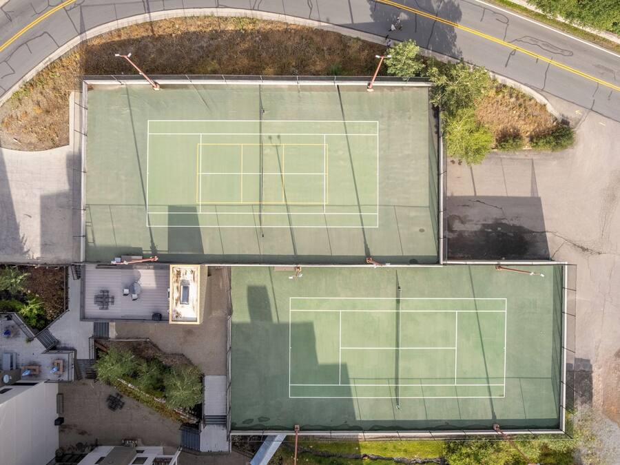 An aerial view of a tennis court complex with two courts, surrounding buildings, and a curved road.