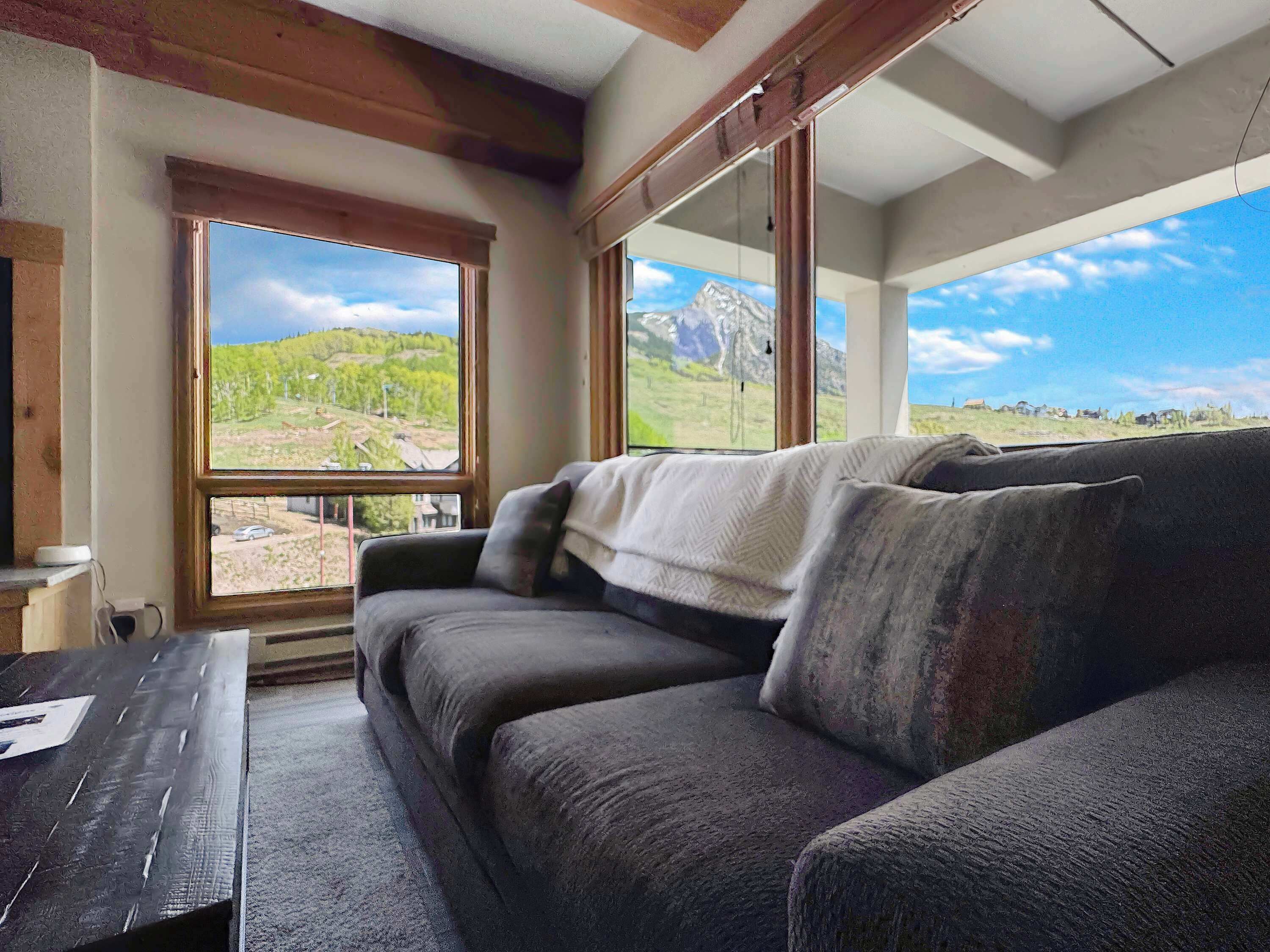 A room with a sofa, a table, and large windows offering a view of a mountainous landscape.