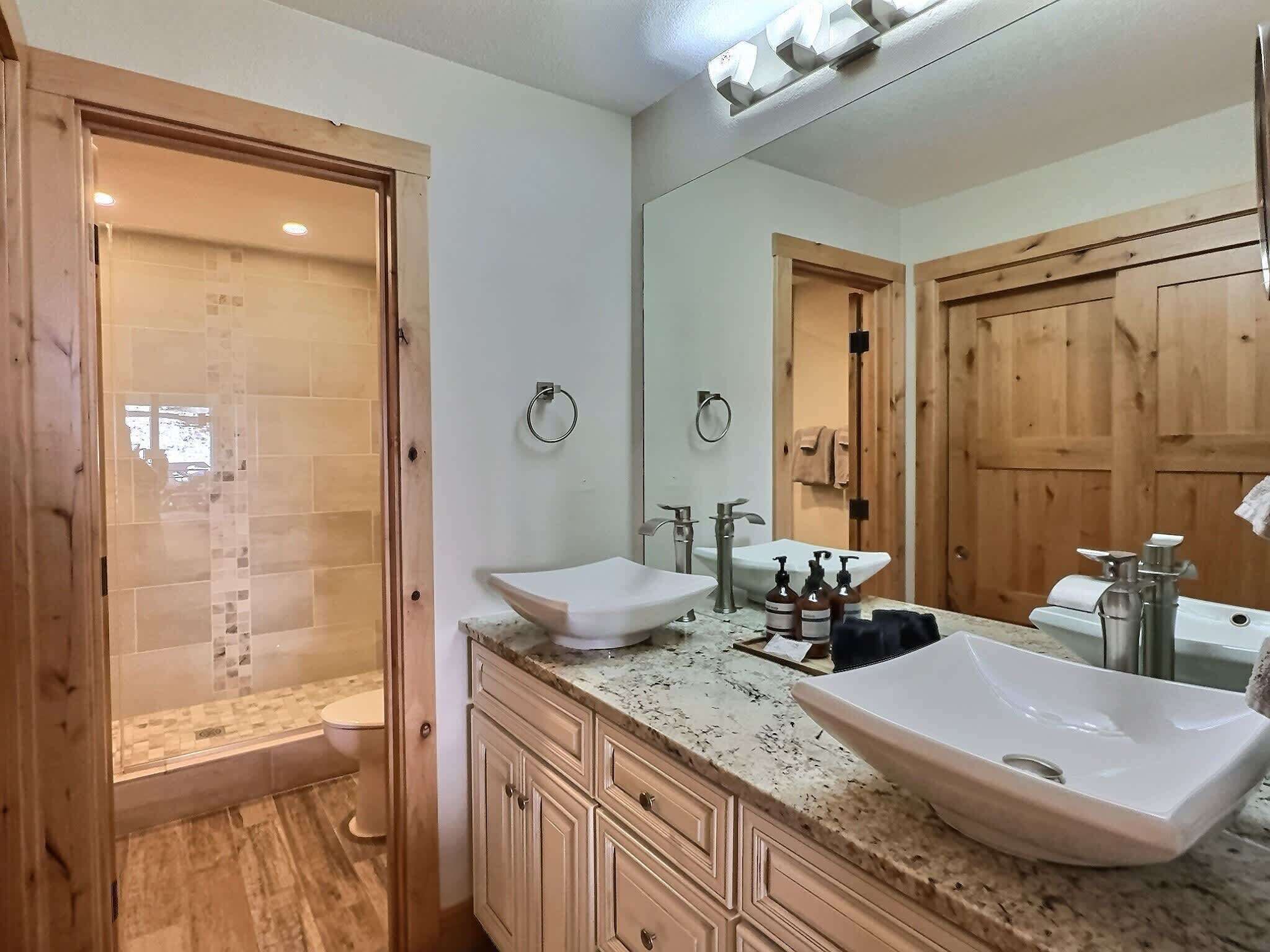 A modern bathroom with a large mirror, two sinks, wooden cabinets, and a shower area.