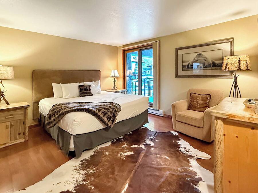 A hotel room with a bed, a chair, a desk, a cowhide rug, a window with a view, and a painting on the wall.