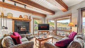 Ein gemütliches Wohnzimmer mit gemusterten Sofas, einem Kamin und einem großen Fernseher, der 'NETFLIX' anzeigt.