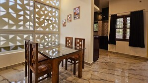 A dining area with a glass table and wooden chairs, a decorative window with geometric patterns, and a kitchen visible through an open door. - Bricknest Hillview (Mysore)