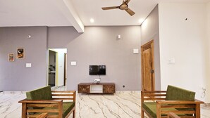 A spacious living room with a marble floor, two green upholstered chairs, a wooden coffee table, a flat-screen TV mounted on the wall, and a ceiling fan. - Bricknest Hillview (Mysore)