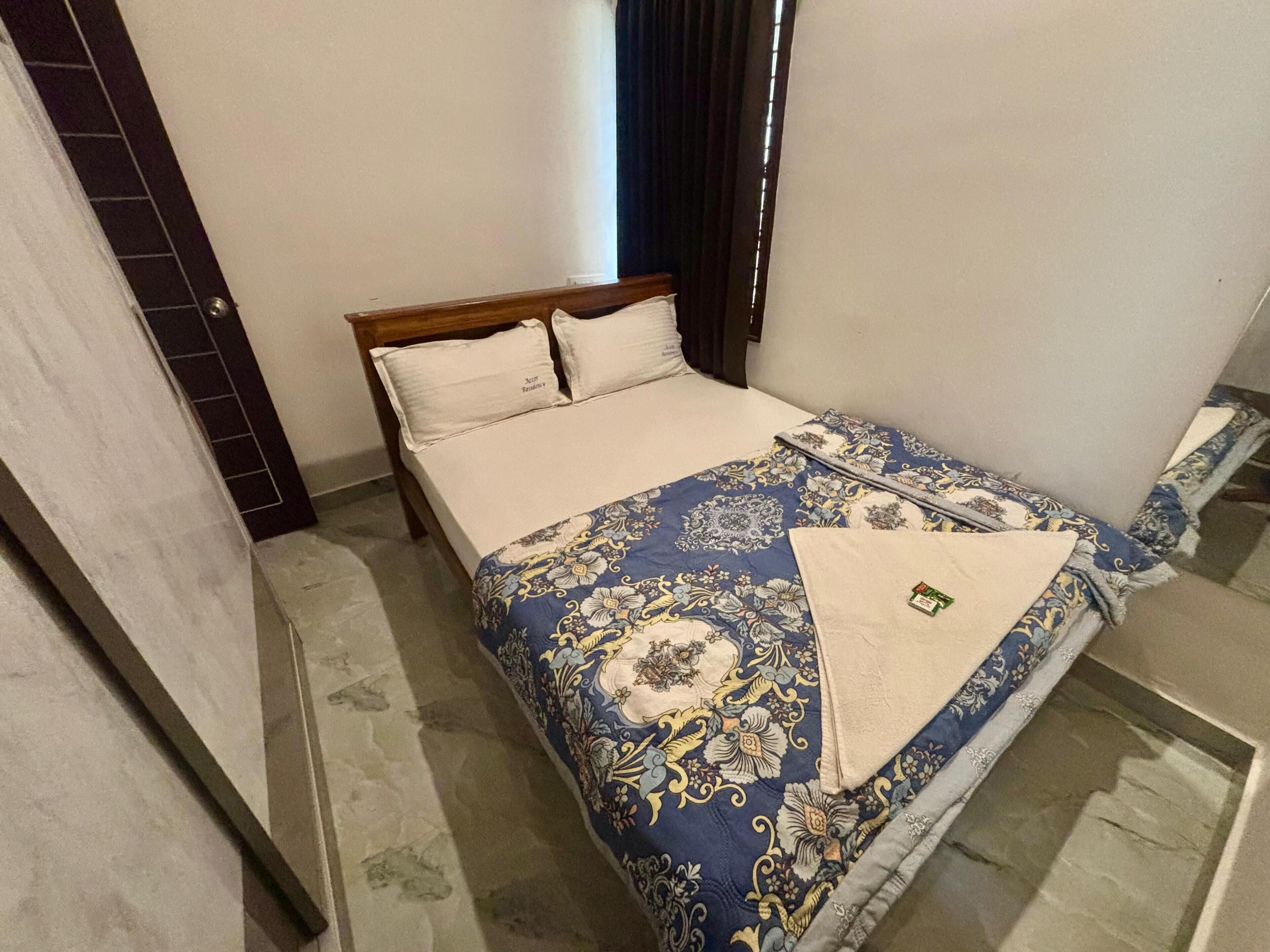 Een net opgemaakt bed met een bloemenprintdeken, een nachtkastje met een lamp en een raam met gordijnen.