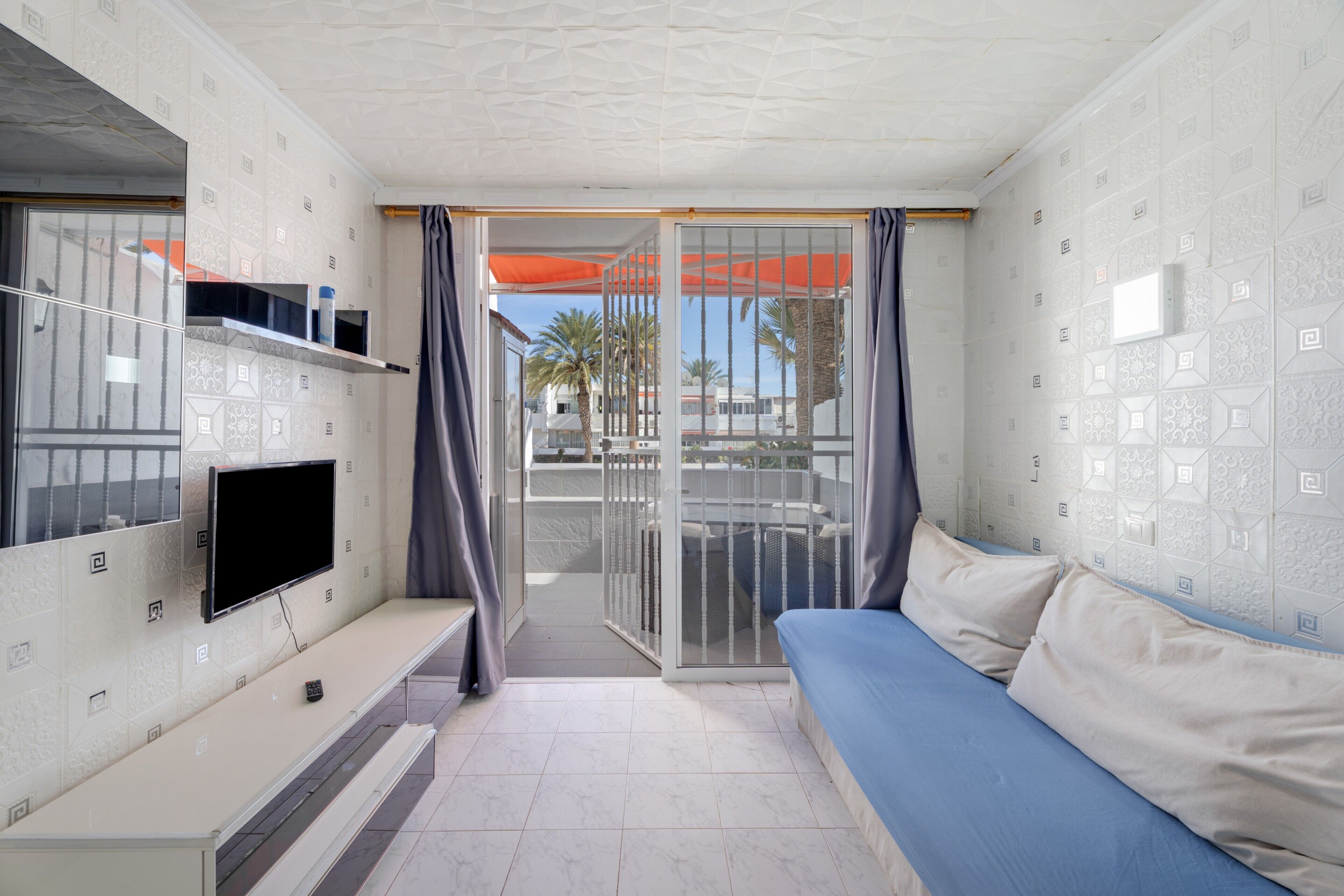 A modern room with a TV, sofa, and a balcony with a view of palm trees.