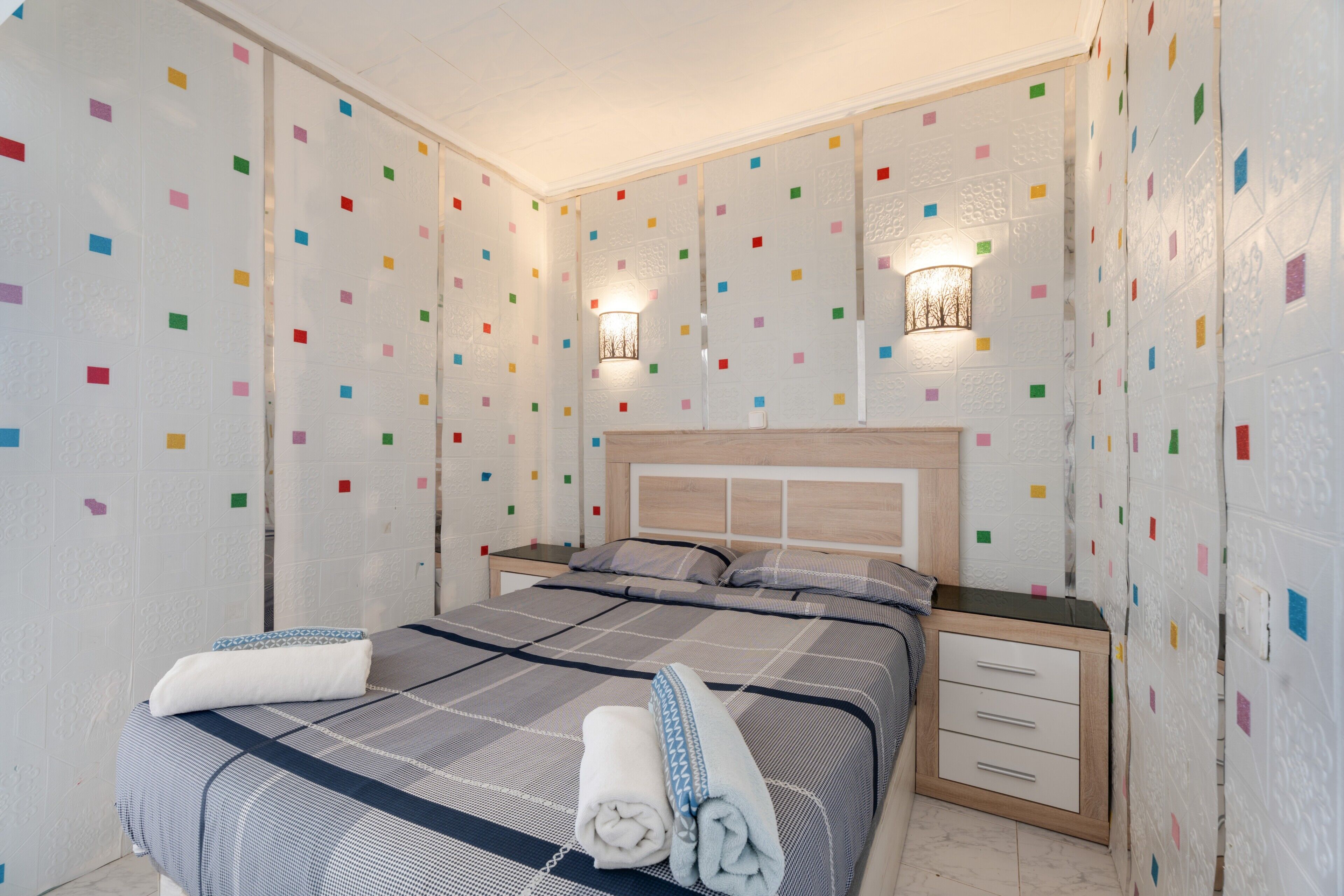 A neatly made bed with a checkered bedspread, two bedside tables with lamps, and a wall with colorful square patterns.