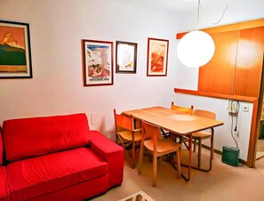 Un espace de vie compact comprenant un canapé rouge, une table à manger en bois avec des chaises, et un coin cuisine avec un meuble blanc et un luminaire suspendu.