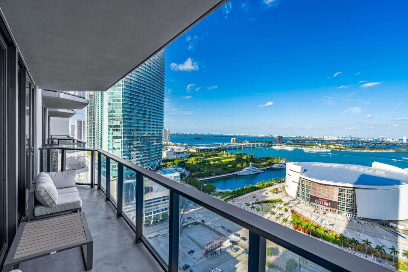 Ein Balkon mit Blick auf eine Marina, ein modernes Gebäude und einen klaren, blauen Himmel.