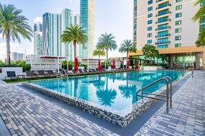 A swimming pool with lounge chairs and umbrellas, surrounded by palm trees and modern buildings. - Lux 2BD Apt: Stunning Water Views (Miami)