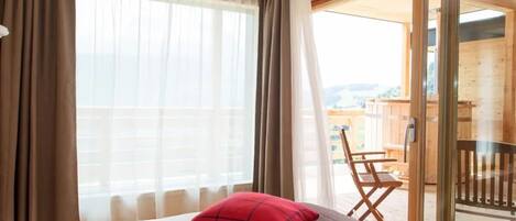 A hotel room with a bed, a red pillow, a wooden chair, and a balcony with a view.