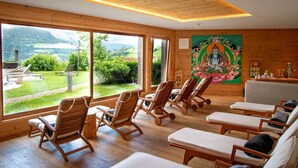 Une pièce aux murs en bois et aux grandes fenêtres, avec des chaises longues et un tableau représentant une divinité.