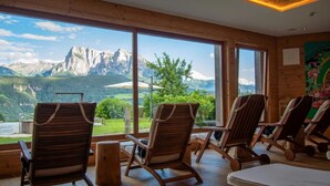 Une pièce aux murs en bois et aux grandes fenêtres donnant sur une vue imprenable sur les montagnes. On y trouve plusieurs chaises en bois et un lit.
