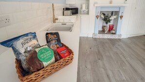 A kitchen with white cabinets, a microwave, and a sink. A wicker basket contains food items like Tesco Finest Lightly Salted Sea Salt, Slater Fyres Plum Bread, and Fox's Chocolatier Chocolate Chunk Cookies. - The Seaside Retreat (Mablethorpe)