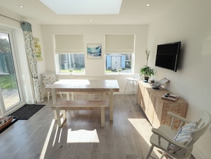 A modern kitchen with a wooden table, a white chair, a TV mounted on the wall, and a window with a view of the outside. - The Seaside Retreat (Mablethorpe)