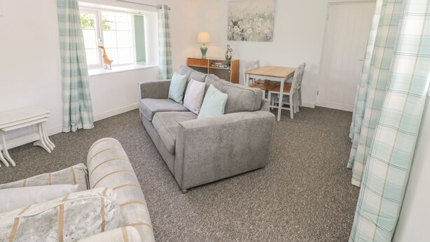 Un salon chaleureux avec un canapé gris, une table à manger avec des chaises, une petite table blanche et une fenêtre avec des rideaux.
