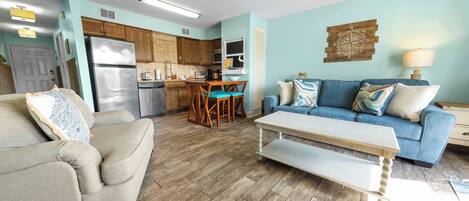 A living room with a sofa, a coffee table, a dining area with chairs, a kitchen with appliances, and a blue wall with a compass decoration.