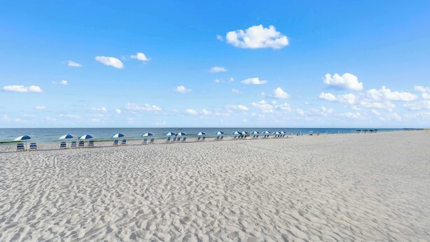 Ampia spiaggia sabbiosa con file di sedie a sdraio e ombrelloni azzurri e bianchi, cielo sereno e oceano sullo sfondo.