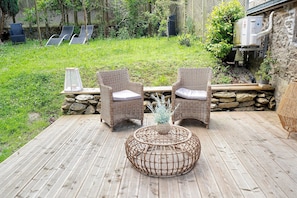 Eine Holzterrasse mit Korbstühlen und einem gewobenen Couchtisch, eine Steinmauer und ein Garten mit Gras und Pflanzen.