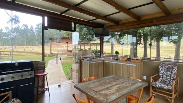 Een terras buiten met een houten tafel en stoelen, een barbecue en uitzicht op een omheind gebied met bomen.