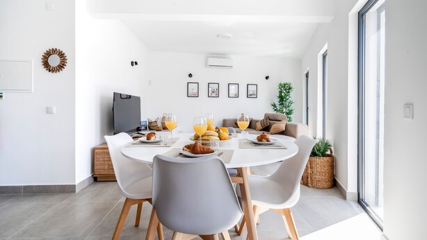 A modern dining area with a white table, grey chairs, and a TV. There are framed pictures on the wall, a plant, and a wicker basket. - Portimao riverview T2 luxe 1st floor with Balcony, Wi-Fi and Air Conditioning (Portimão)