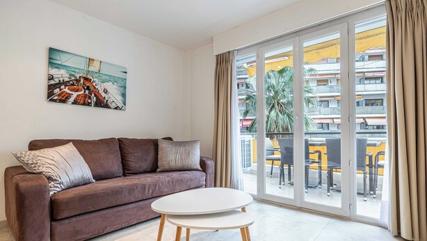 Un salón moderno con un sofá marrón, una mesa redonda blanca y un gran ventanal que da a un balcón exterior con sillas amarillas y vistas a edificios.