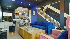 Un salon moderne avec un canapé bleu, une table basse en bois et un fauteuil rose. On aperçoit, à l’arrière-plan, un escalier orné d’un motif géométrique sur le mur.