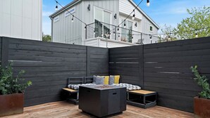 Un espace de détente extérieur moderne, doté d’une terrasse en bois, d’un canapé noir central et de deux petits bancs.