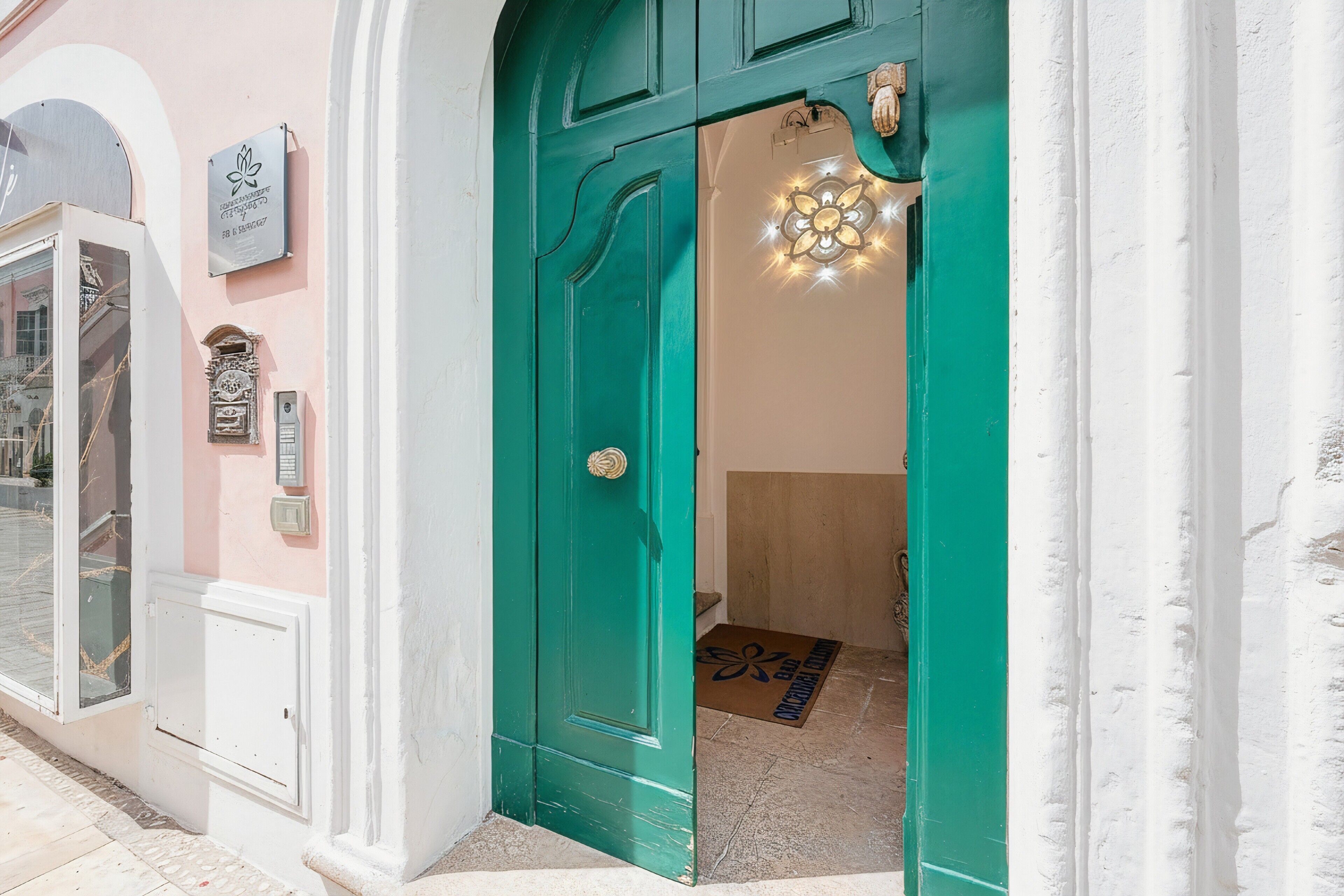 Una porta color petrolio con un portalampada sopra, un tappetino d'ingresso e un cartello fissato al muro.