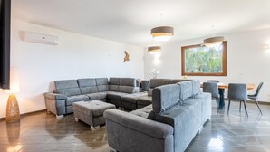 A modern living room with a sectional sofa, a dining table with chairs, pendant lighting, and a large window with a view of greenery. - Villa Altamarea - Scopello (Scopello)