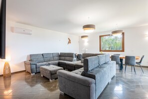 A modern living room with a sectional sofa, a dining table with chairs, pendant lighting, and a large window with a view of greenery. - Villa Altamarea - Scopello (Scopello)