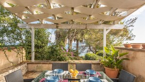 Un espace extérieur aménagé pour les repas, avec une pergola, des chaises en osier, une table en verre et une vue sur la mer.