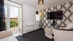 Ein Zimmer mit einer Couch, einem Sessel, einem an der Wand befestigten Fernseher, einer Heizung und einem Fenster mit Blick ins Grüne.