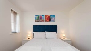 Ein Schlafzimmer mit einem großen Bett, zwei Nachttischen mit Lampen, einem Fenster mit Jalousien und drei an der Wand hängenden Bildern.