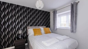Un dormitorio con papel tapiz estampado, una cama con ropa de cama blanca y dos almohadas amarillas, una mesita de noche con una lámpara, y una ventana con cortinas.