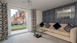 Una sala de estar con sofá beige, cojines estampados, una mesa de centro de vidrio y vista a un jardín trasero a través de una puerta corrediza de vidrio.