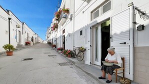 Een smalle straat met witte gebouwen, een vrouw die op een bankje voor een winkel zit, en een kat die er in de buurt ligt.
