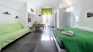 Een moderne hotelkamer met een groene bank, een bed met een groene dekbedovertrek, een zithoek met een tafel en stoelen, en uitzicht op de straat door het raam.