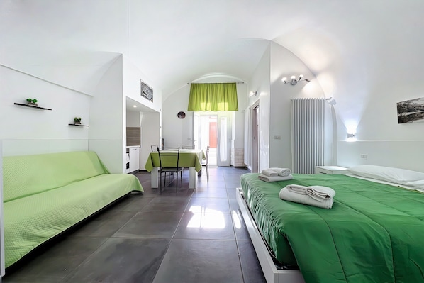 Una habitación de hotel moderna con un sofá verde, una cama con edredón verde, un área de comedor con mesa y sillas, y vista a la calle por la ventana.