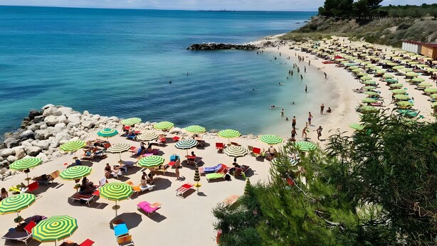 Een strand met veel parasols en ligbedden, een rotsachtige kustlijn en een helderblauwe lucht.