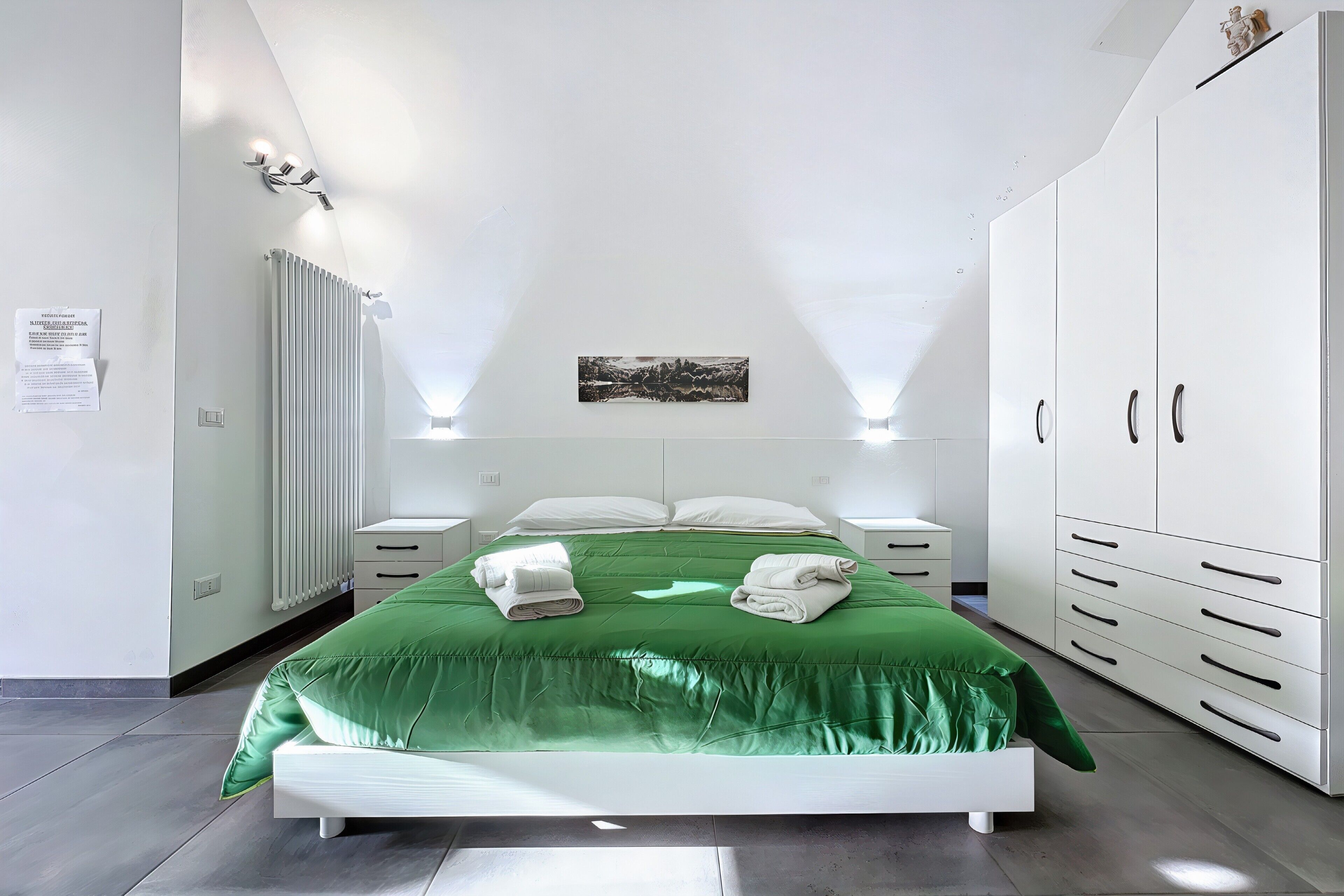 Une chambre à coucher moderne avec une couvre-lit verte, du linge de lit blanc, une armoire blanche et un luminaire au plafond.