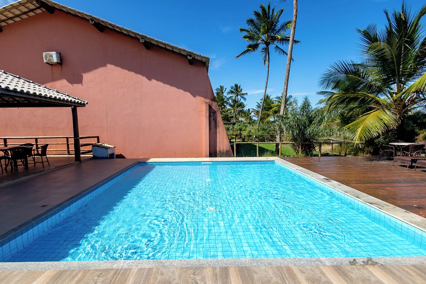 A swimming pool with a tiled deck, a covered seating area, and palm trees in the background.