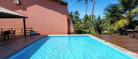 A swimming pool with a tiled deck, a covered seating area, and palm trees in the background.