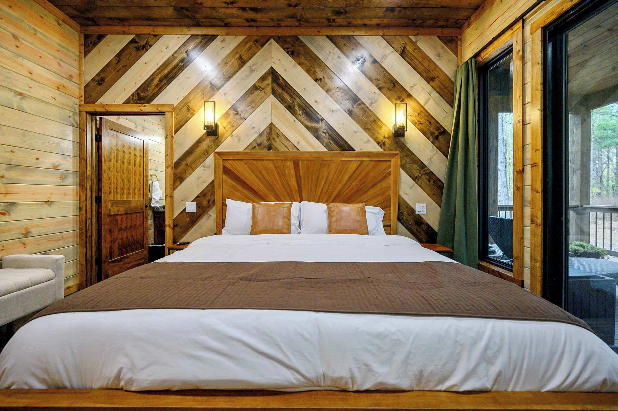 Un dormitorio de cabaña de madera con una cama grande, dos lámparas fijadas a la pared y vistas del exterior a través de una ventana.