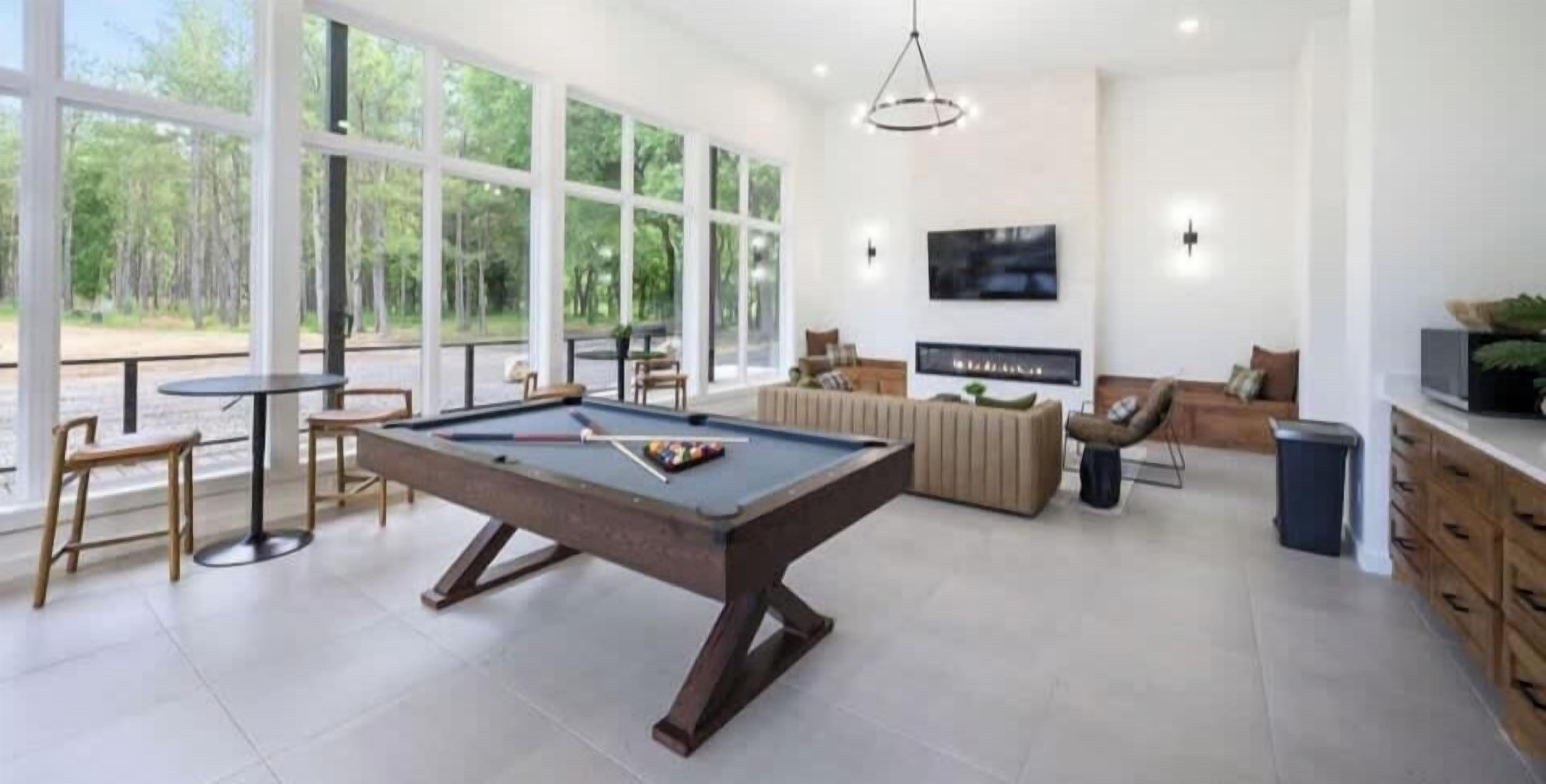 A modern living room with a pool table, comfortable seating, a flat-screen TV, and large windows offering a view of the outdoors.