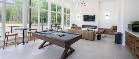 A modern living room with a pool table, comfortable seating, a flat-screen TV, and large windows offering a view of the outdoors.