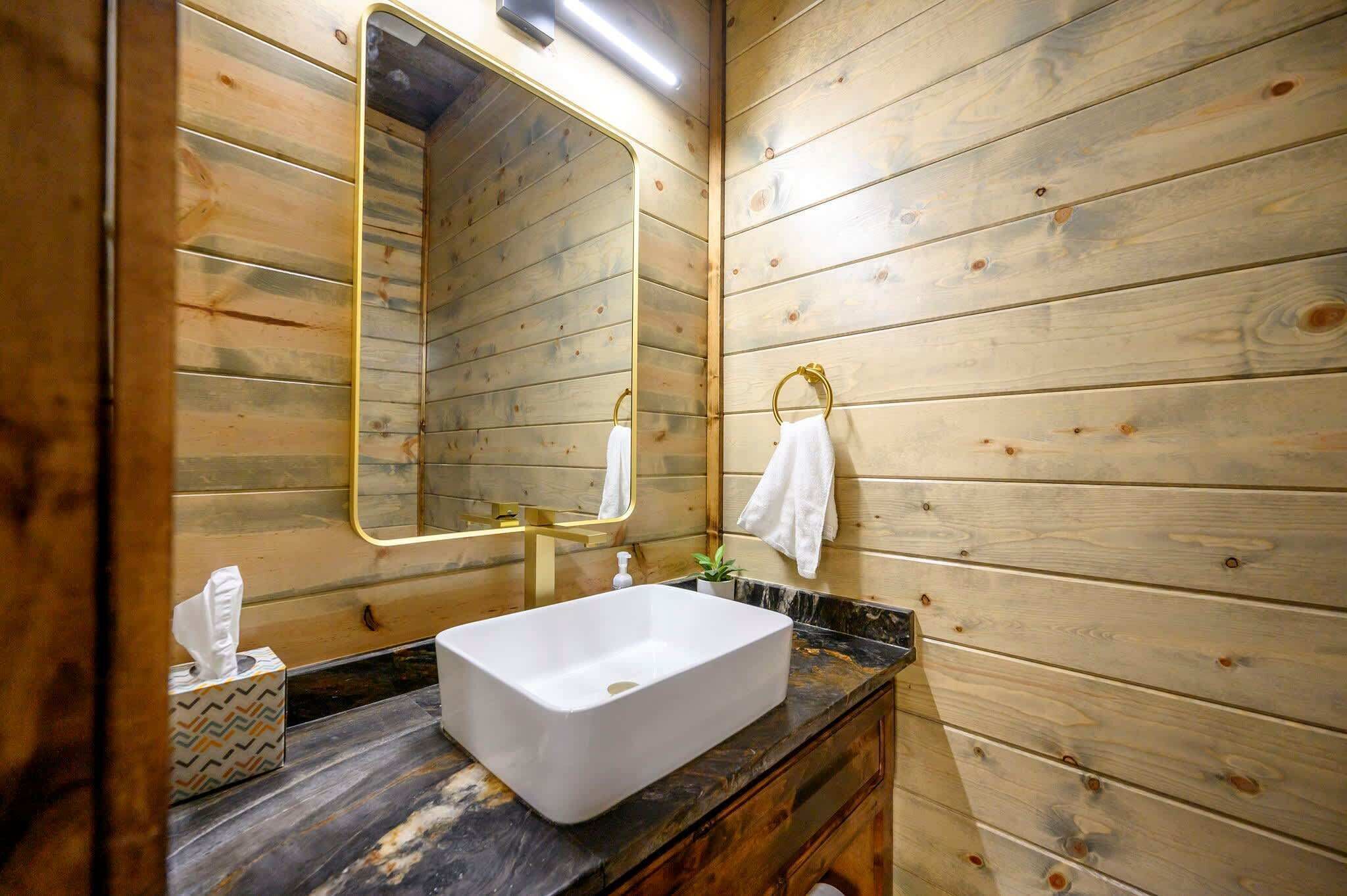 A wooden-paneled bathroom with a rectangular sink, a gold-framed mirror, and a towel hanging on the wall.