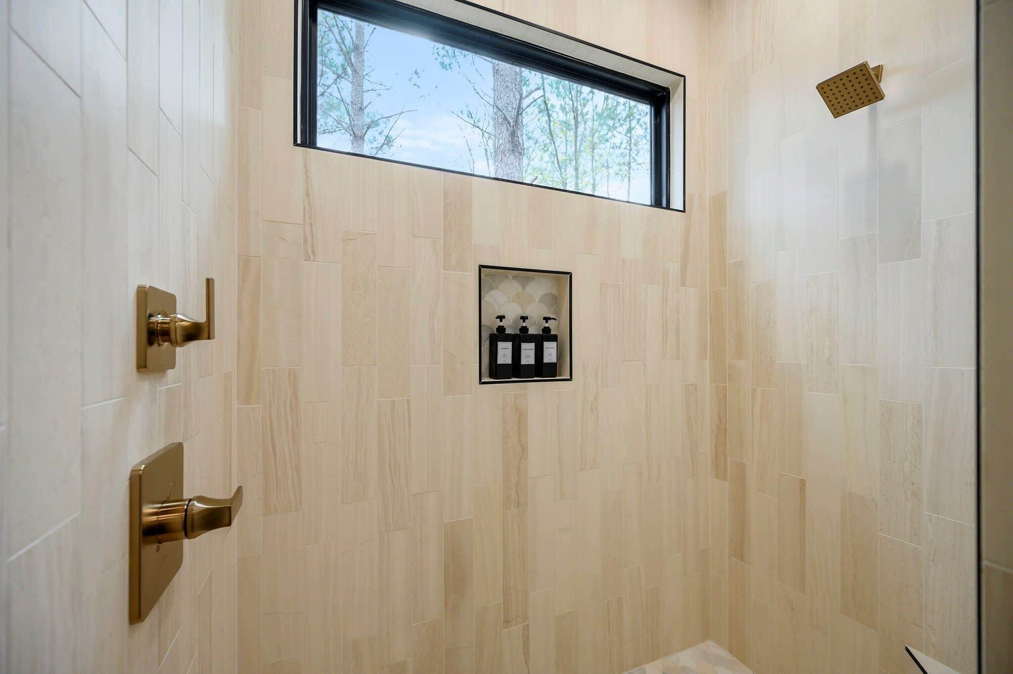 Een moderne douche met een raam erboven, betegelde wanden en een klein plankje met toiletartikelen.