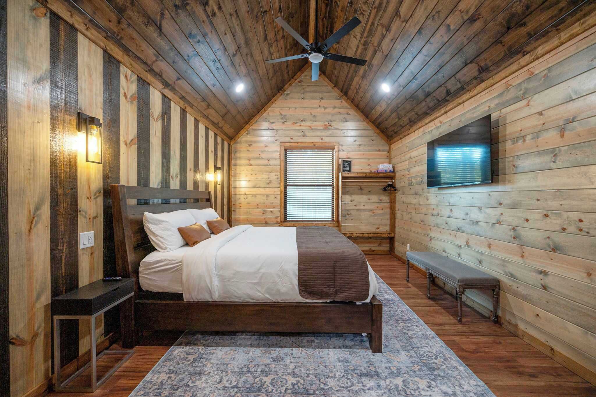 A cozy bedroom with a wooden ceiling, a bed, a bench, and a TV mounted on the wall.