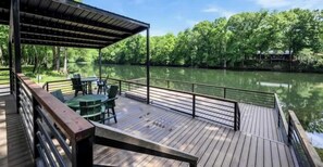 Une terrasse en bois avec une table et des chaises, donnant sur un lac paisible et entourée d’arbres.