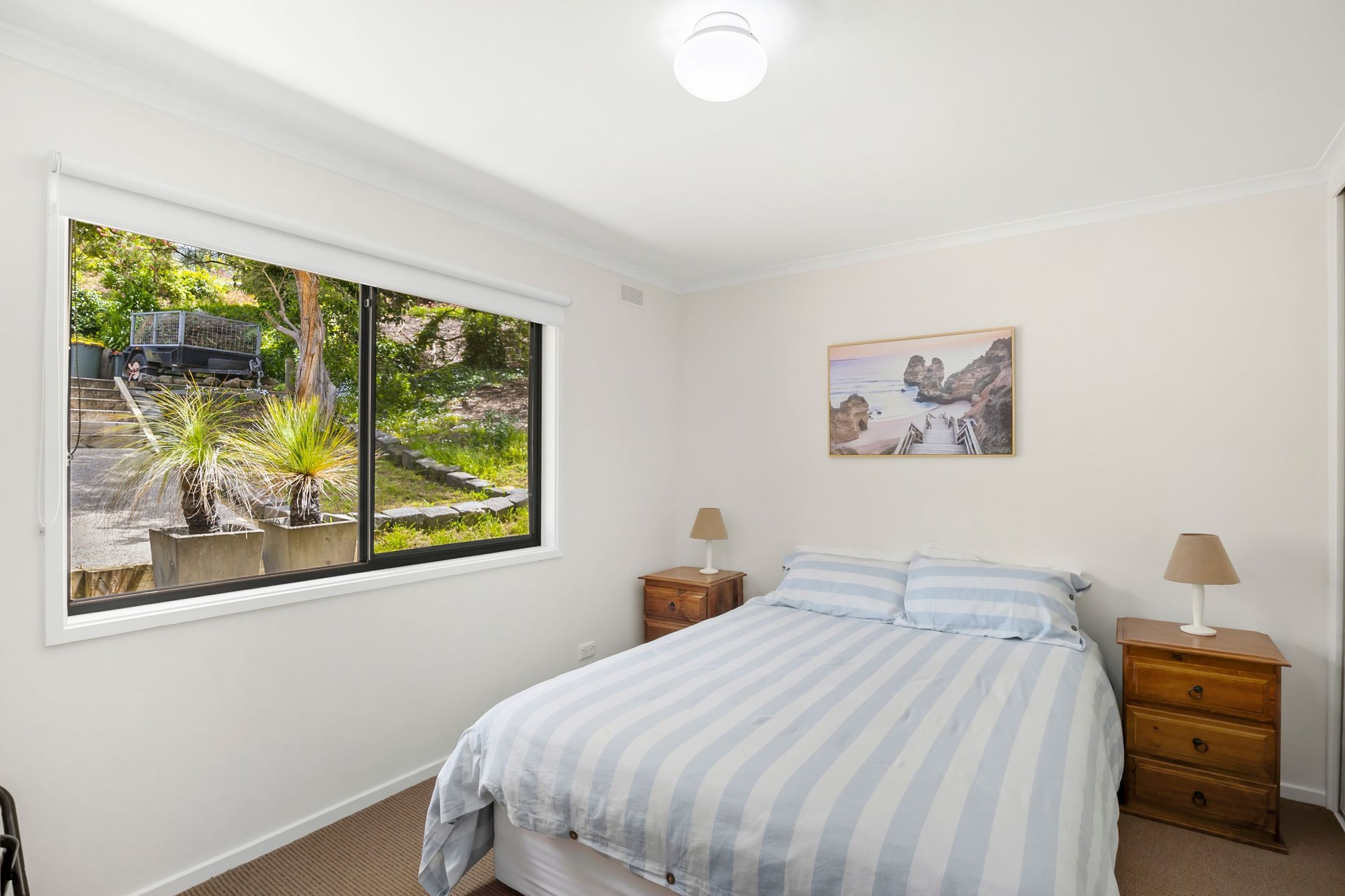A bedroom with a bed, two bedside tables, a window with a view of a garden and a parked vehicle, and a painting on the wall.